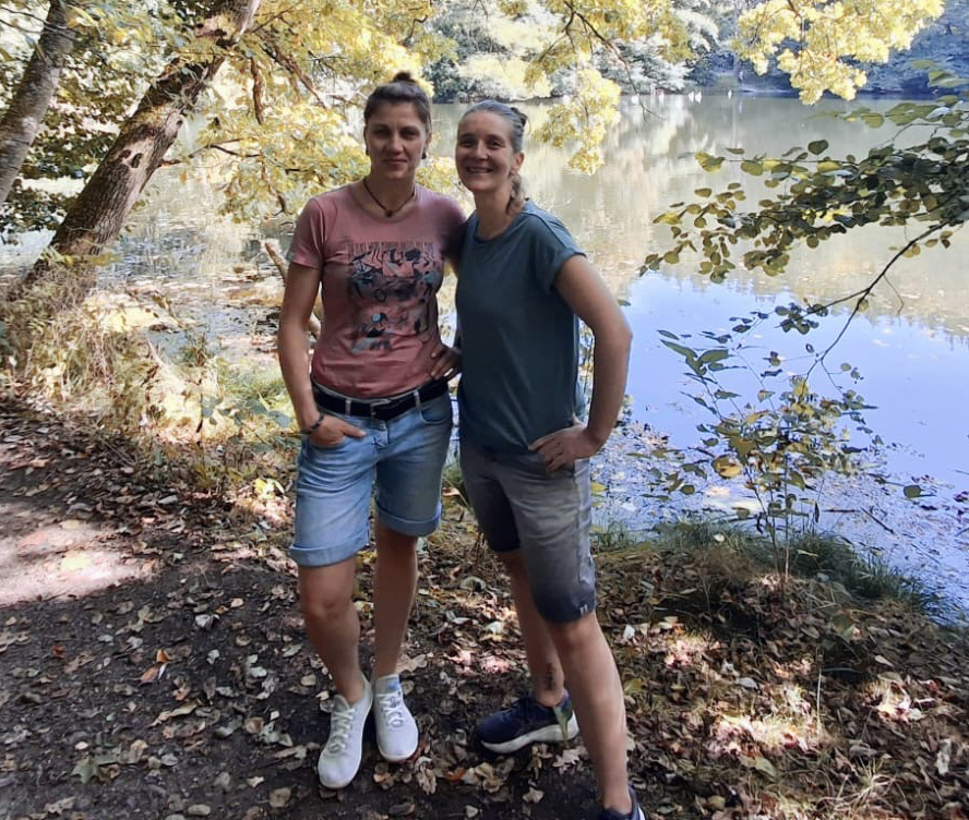 Claudia und Lydia stehen im Wald vor einem Gewässer